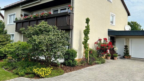 Un immeuble résidentiel à deux étages, doté d’un balcon, d’un garage et d’un jardin bien entretenu où poussent diverses plantes et fleurs.