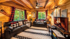 A cozy cabin interior with a fireplace, leather sofas, and a large window overlooking greenery. - Chalet in the Trees - Sauna|Game Room|Pool Access (Bellaire)