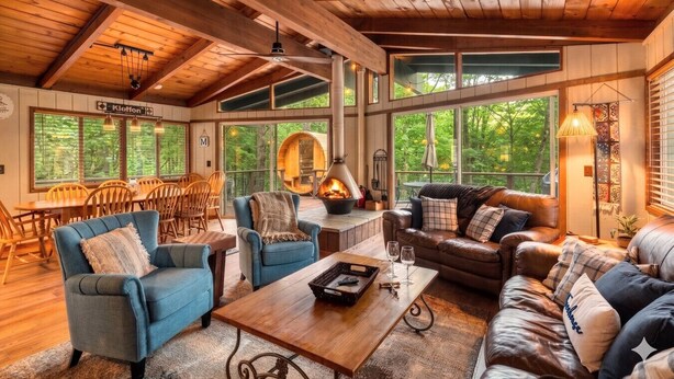 A cozy cabin interior with wooden beams, a fireplace, and large windows offering a view of the forest. - Chalet in the Trees - Sauna|Game Room|Pool Access (Bellaire)