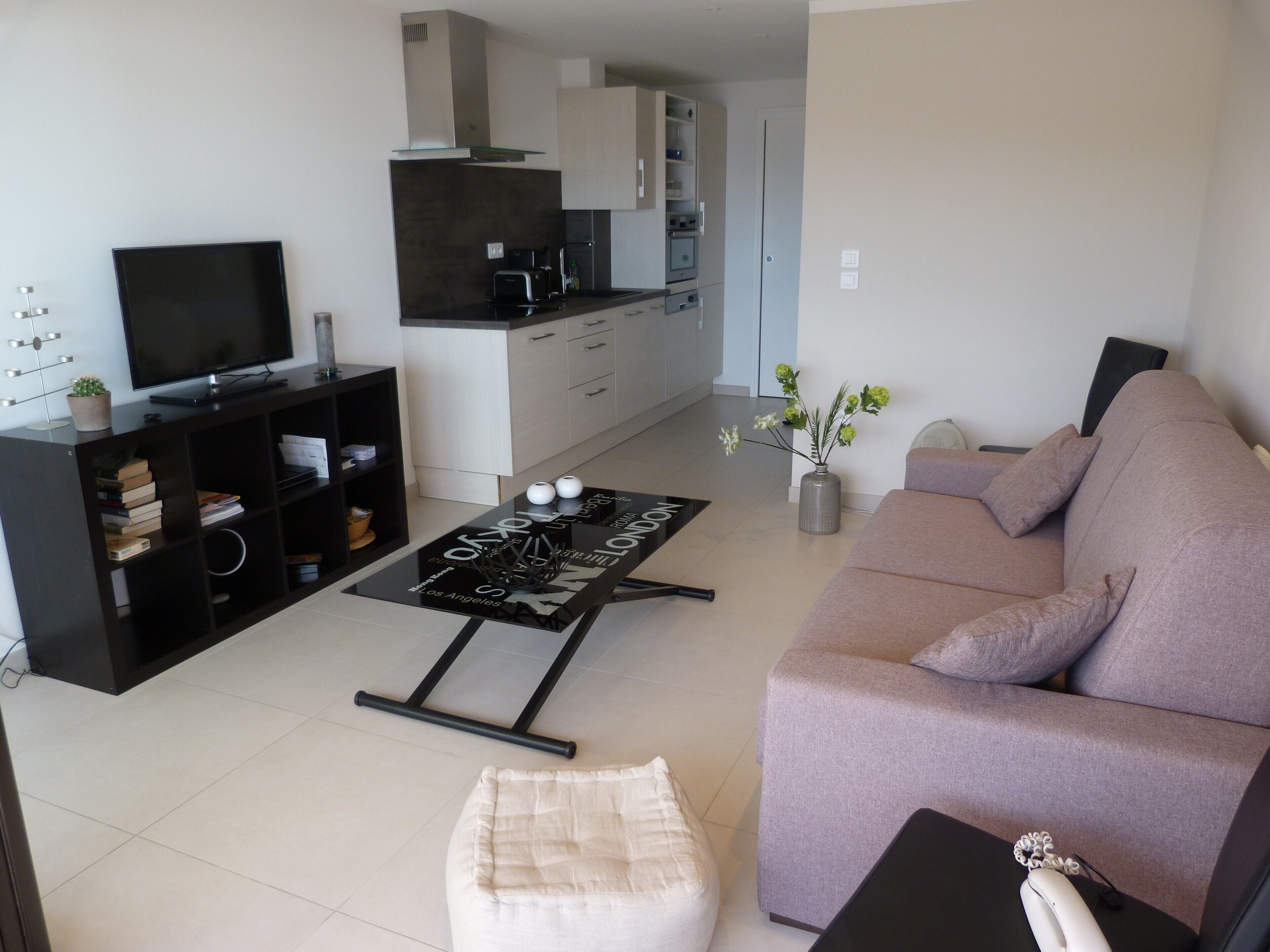 A modern living room with a flat-screen TV, a sofa, a coffee table, and a kitchen area in the background.