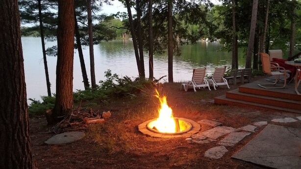 A fire pit surrounded by stone steps, overlooking a lake with chairs and a table on a deck. - Beachfront 1-bedroom home, 15-min. drive to Traverse City.  Private and peaceful (Traverse City)