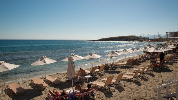 Een strand met ligbedden en parasols, helderblauw water en een glooiend landschap op de achtergrond.