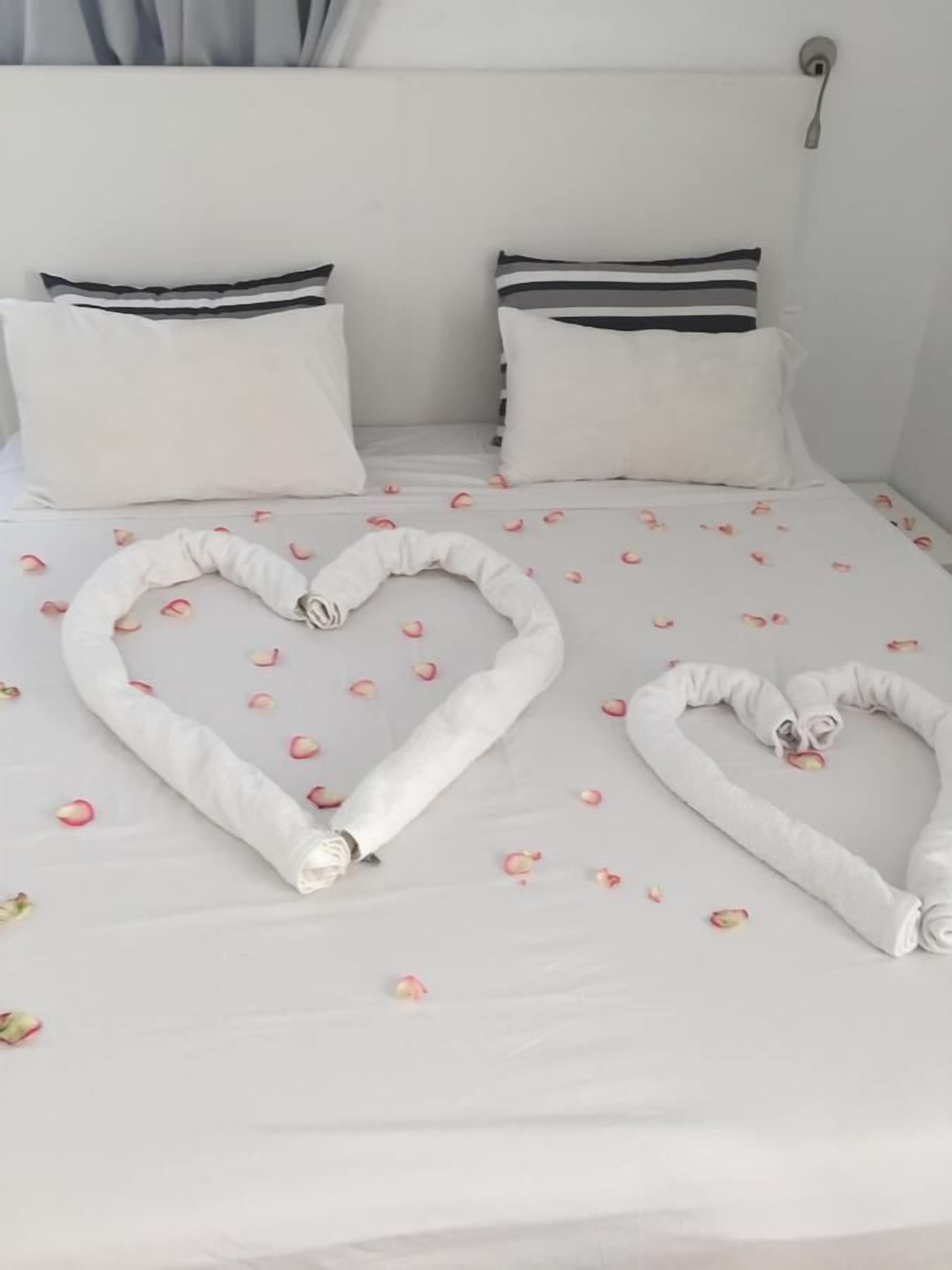 Una cama con sábanas blancas, dos almohadas y toallas con forma de corazón hechas con toallas, adornada con pétalos de rosa.
