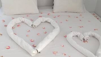 Una cama con sábanas blancas, dos almohadas y toallas con forma de corazón hechas con toallas, adornada con pétalos de rosa.