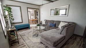 Un salon avec un canapé gris, un banc bleu, une table basse en verre et un tapis à motifs.