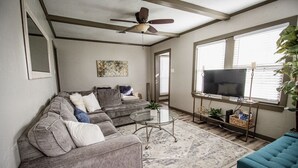 Un salon avec un canapé gris, un fauteuil bleu, une table basse en verre, un ventilateur de plafond, un téléviseur posé sur un meuble et une grande fenêtre avec des stores.