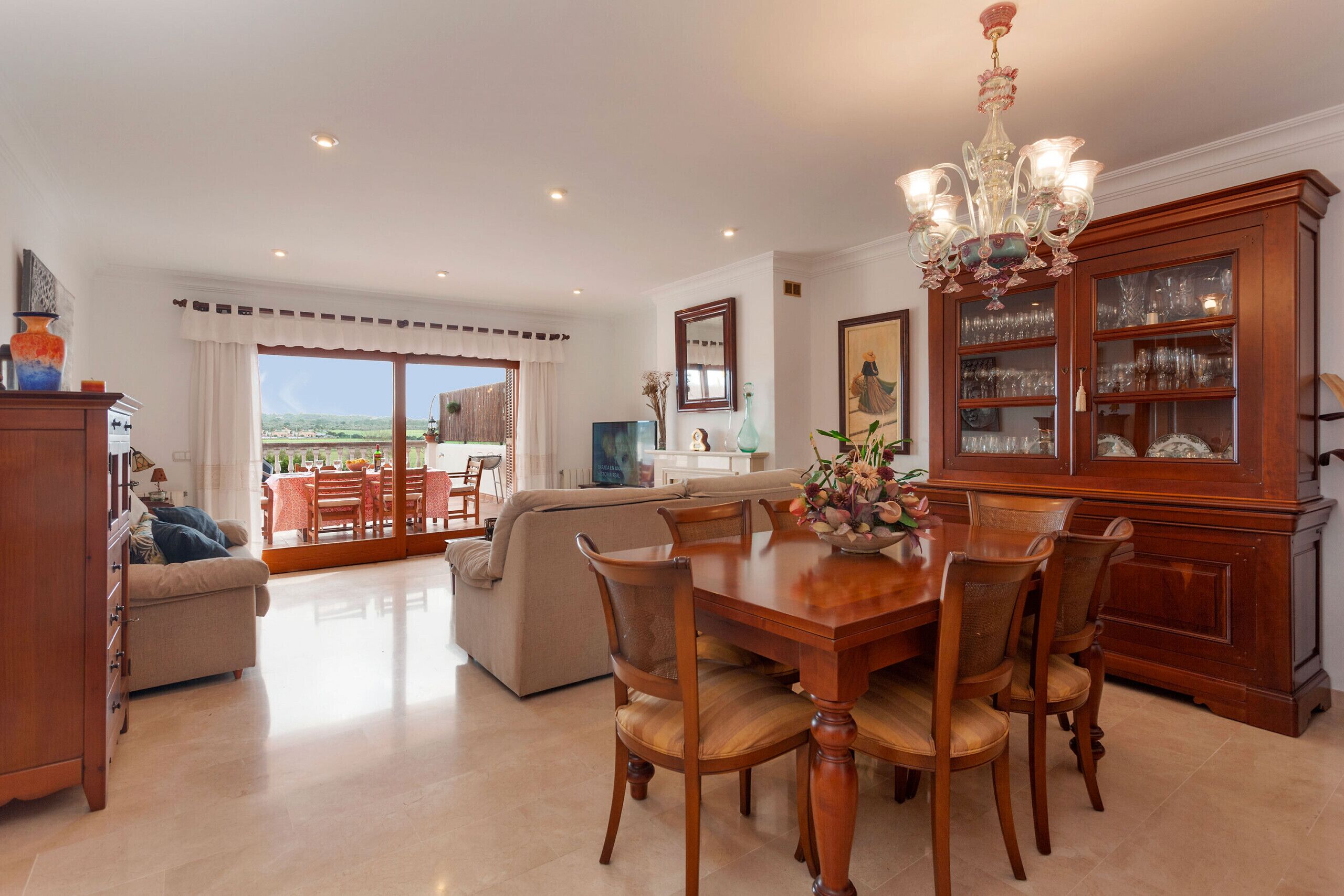 Amplia sala de estar con una mesa de comedor, sillas y una araña. Cuenta con un sofá y un aparador con puertas de vidrio.