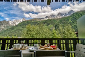 A table set with cups, a teapot, plates, a baguette, and fruit on a balcony with a mountain view. - Stylish Apartment next to Brevent Lift Station (Chamonix-Mont-Blanc)
