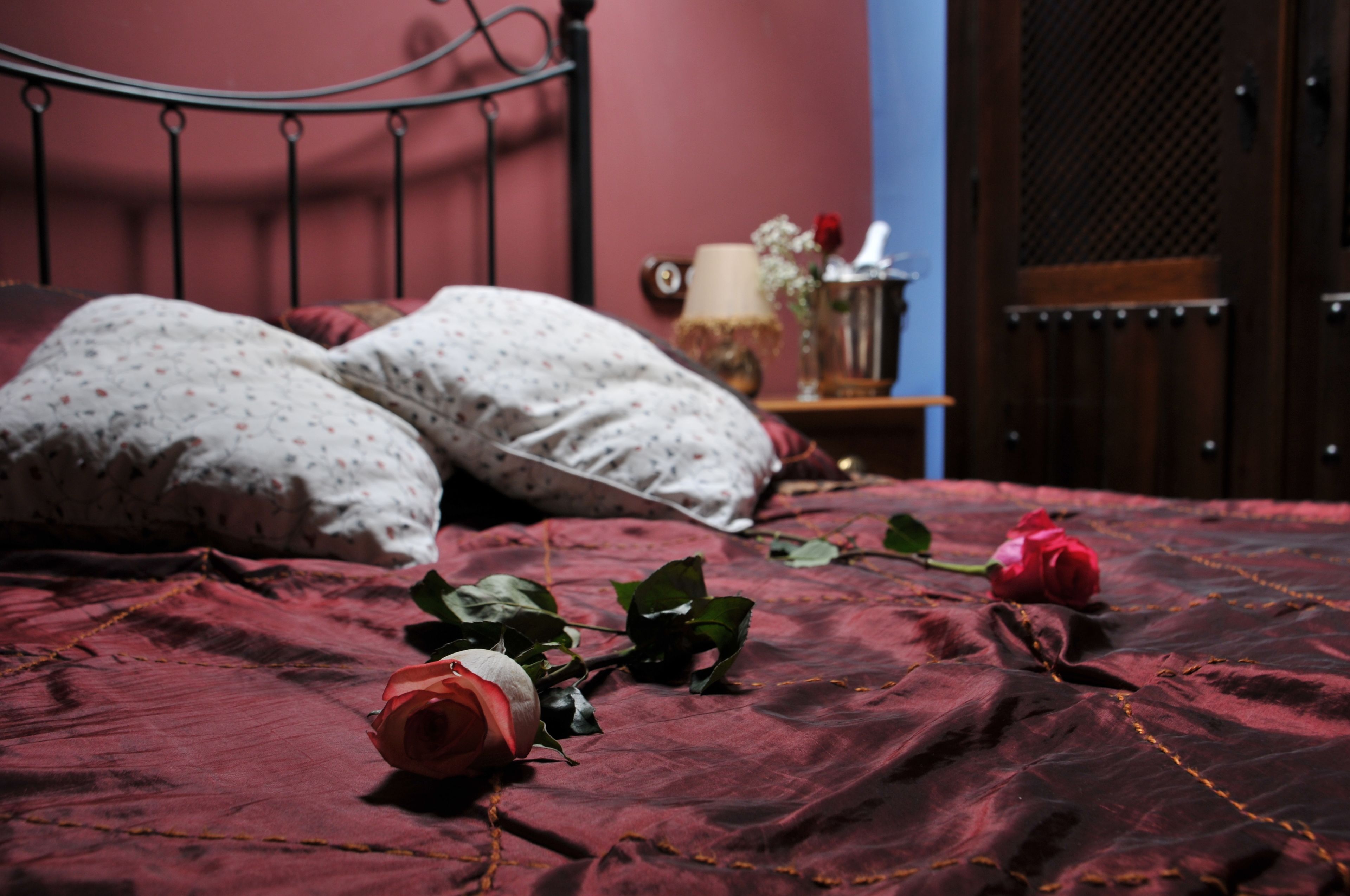 Una cama con una colcha roja, dos almohadas y un florero con flores sobre una mesita de noche.