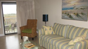 Una sala de estar con un sofá a rayas, una silla de mimbre, una mesa de centro de vidrio y un cuadro con una escena de playa.