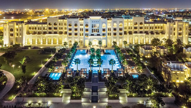 A large, illuminated hotel with a central swimming pool, surrounded by palm trees and a well-maintained lawn.