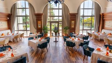 A spacious dining area with multiple tables and chairs, large windows, and a chandelier.