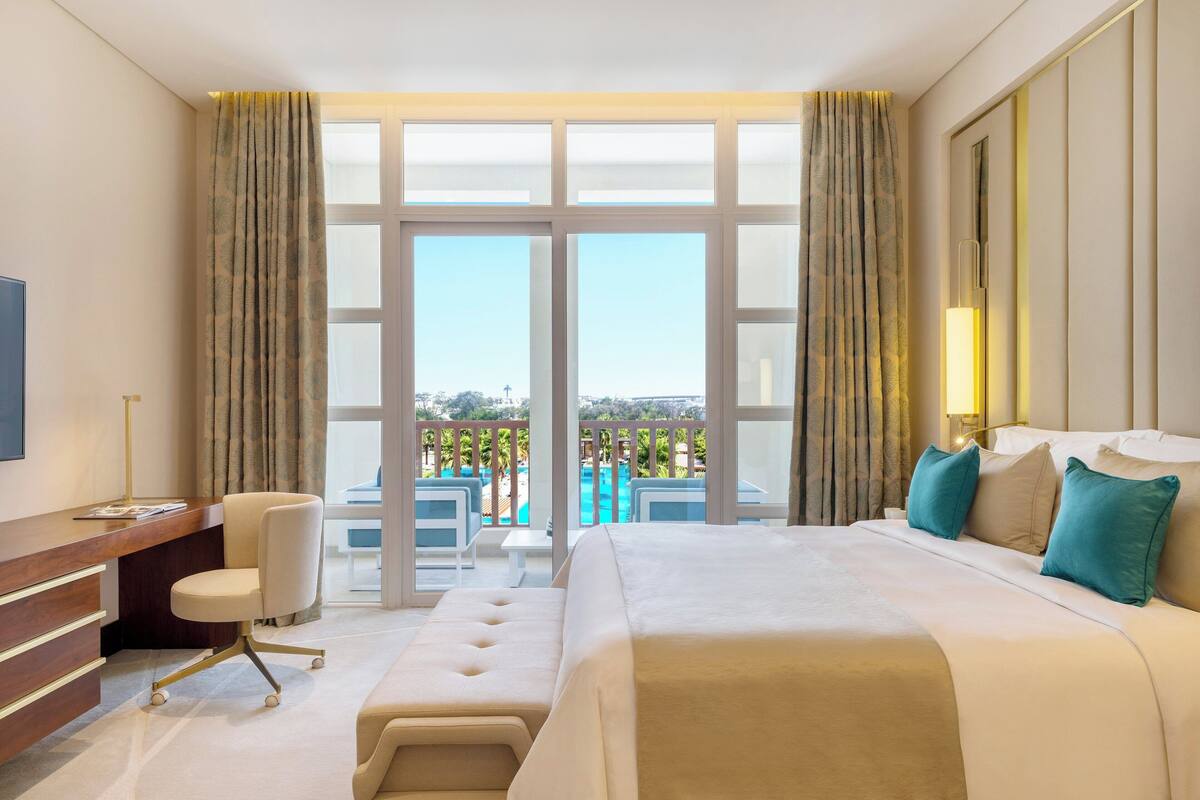 Quarto de hotel com uma cama grande, uma escrivaninha, uma cadeira e vista para a piscina e área externa.
