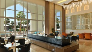 A spacious hotel lobby with a central bar area, seating arrangements, and large windows offering views of palm trees and outdoor seating.