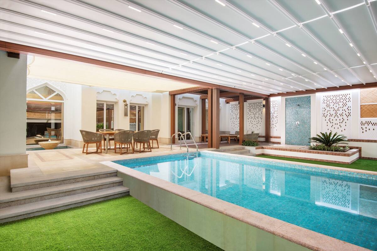 Piscina com degraus que levam a um espaço de pátio, cercada por móveis de madeira e um muro decorativo.
