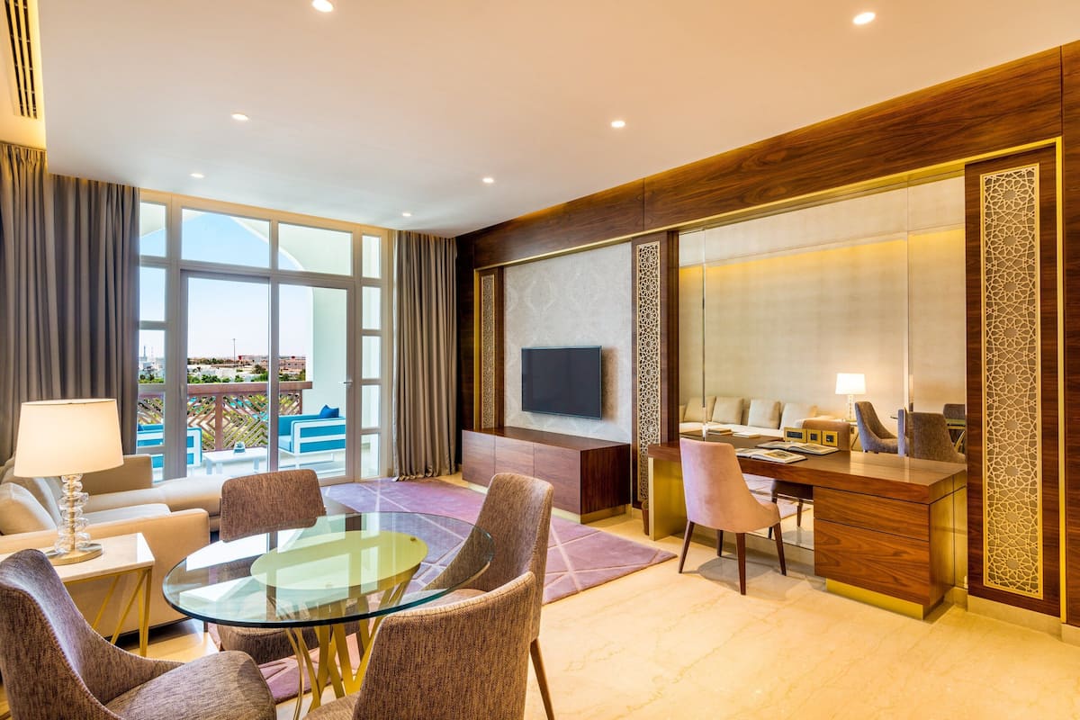 Quarto de hotel moderno com mesa de jantar de vidro, assentos confortáveis, escrivaninha com computador, televisão de tela plana e grandes janelas com vista para o exterior.