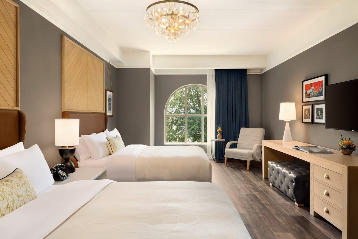 A hotel room with two beds, a desk, a chair, a television, and a large window with a view of trees.