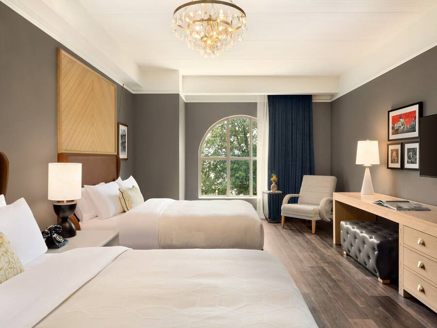 A hotel room with two beds, a desk, a chair, a television, and a large window with a view of trees.