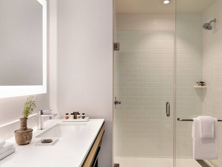 Un baño moderno con ducha de azulejos blancos, cabina de ducha con mampara de vidrio, lavamanos blanco con un florero y toallero con una toalla blanca.