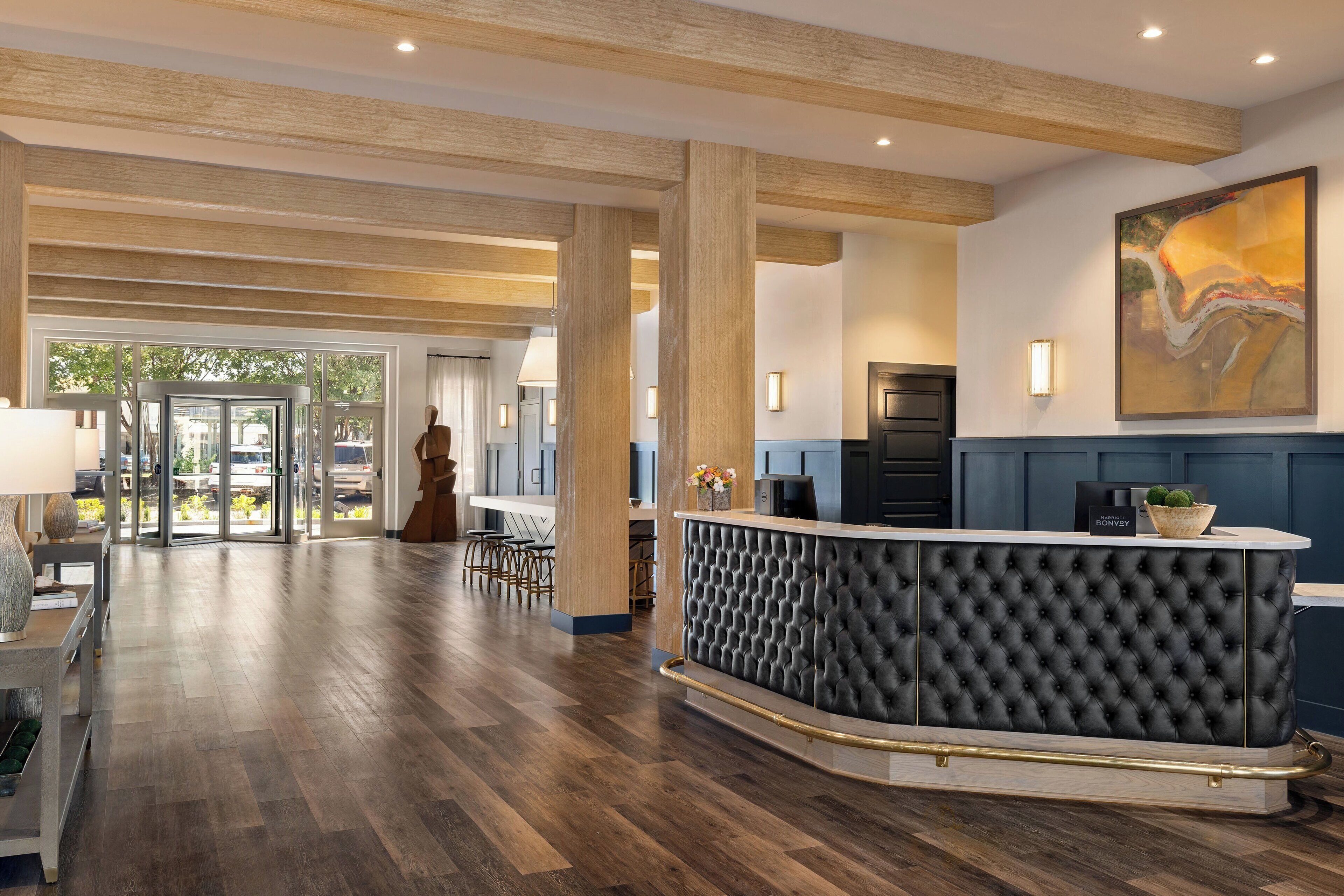 Um hall de hotel com um balcão de recepção preto com tufos, piso de madeira e um grande quadro abstrato na parede.