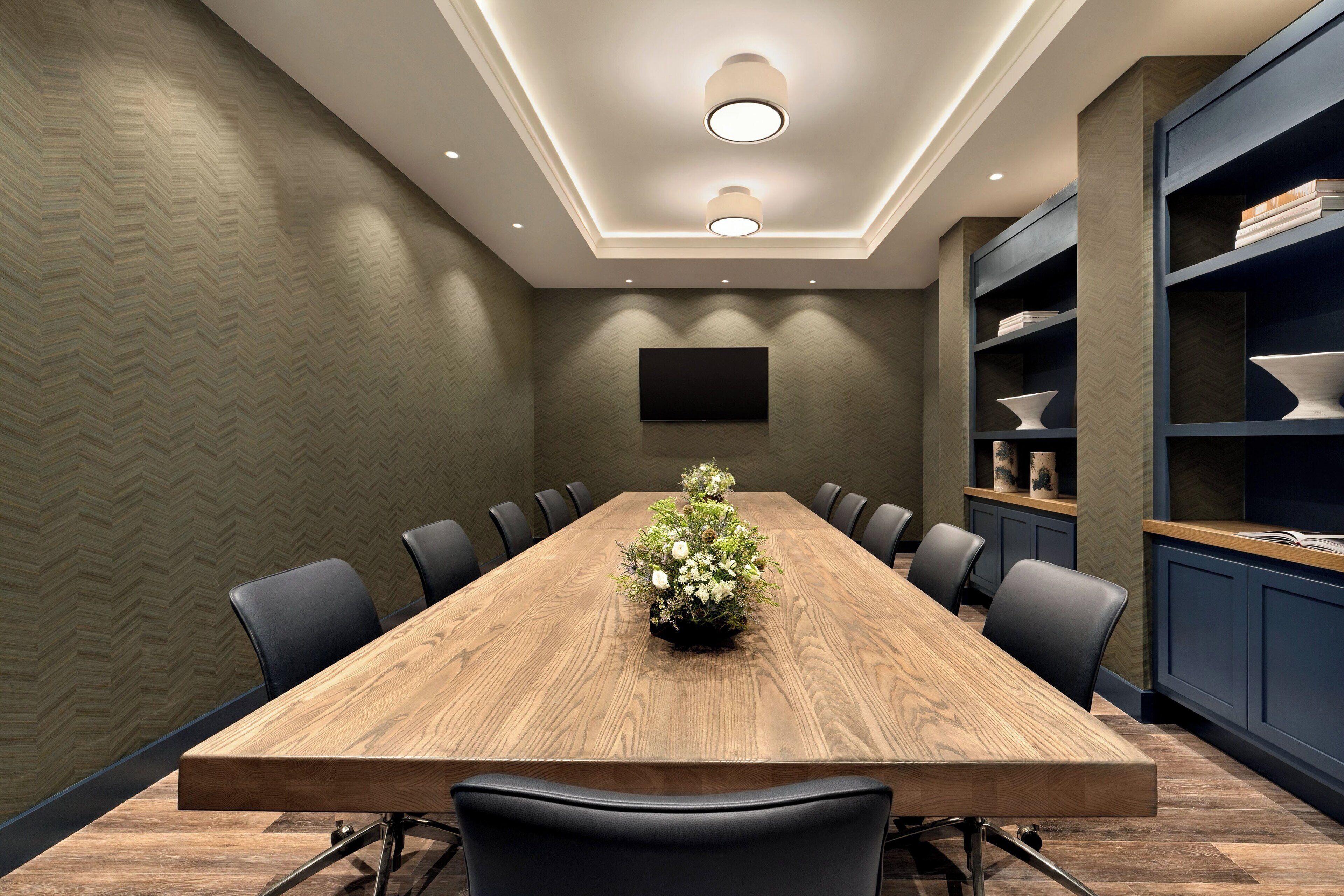 A conference room with a long wooden table, black chairs, a floral centerpiece, a flat-screen TV, and built-in shelving with decorative items.