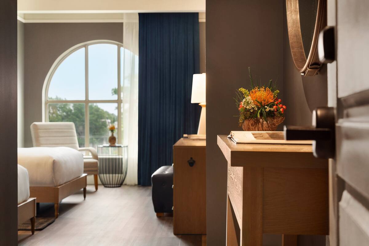 A hotel room with a bed, a desk, a chair, a small table with a lamp, a vase with flowers, and a large window with curtains.