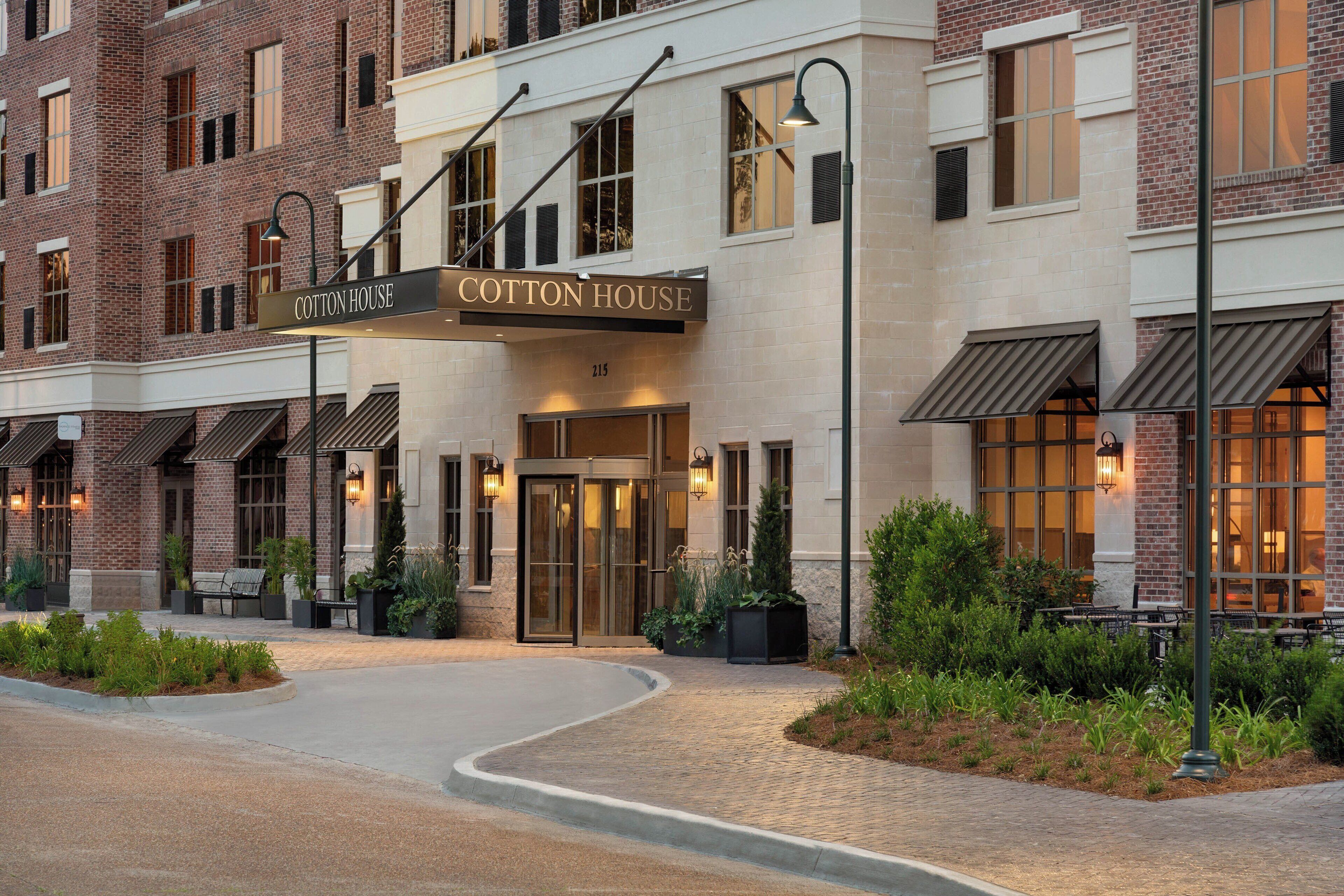 A hotel entrance with a sign reading "Cotton House," featuring a brick facade, a curved driveway, and outdoor seating areas.