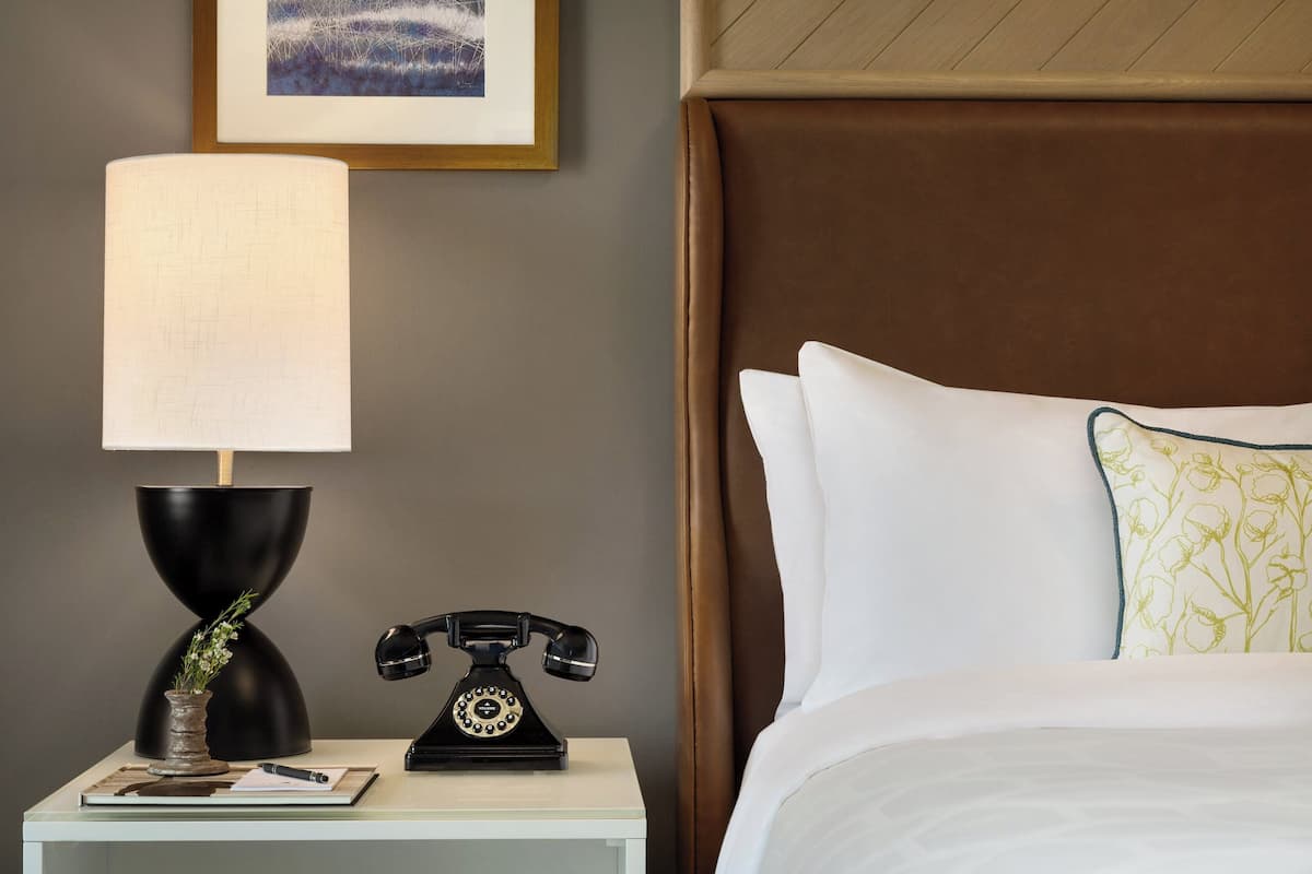 A neatly made bed with white and green pillows, a black telephone, a lamp, and a framed picture on a bedside table.
