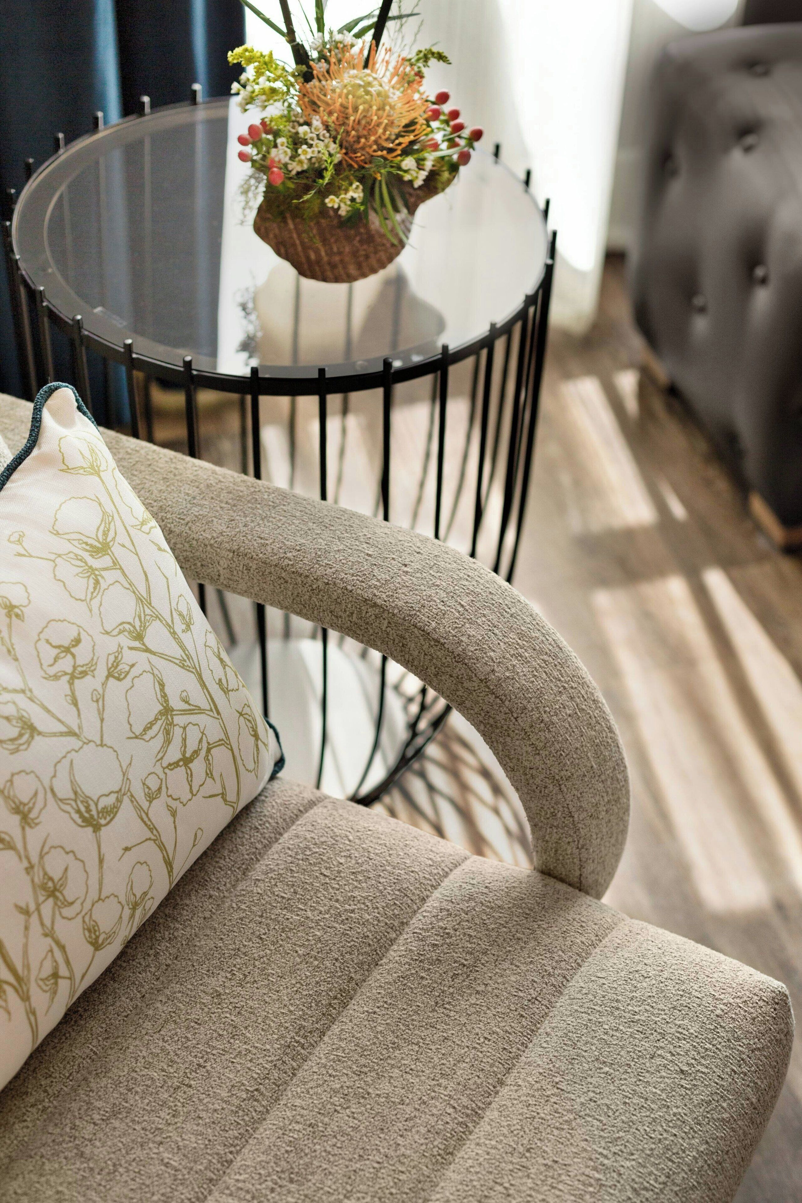 Une table ronde en verre avec un cadre en métal noir, une chaise rembourrée de couleur beige ornée d’un coussin à motifs floraux, et un canapé gris à capitons en arrière-plan.