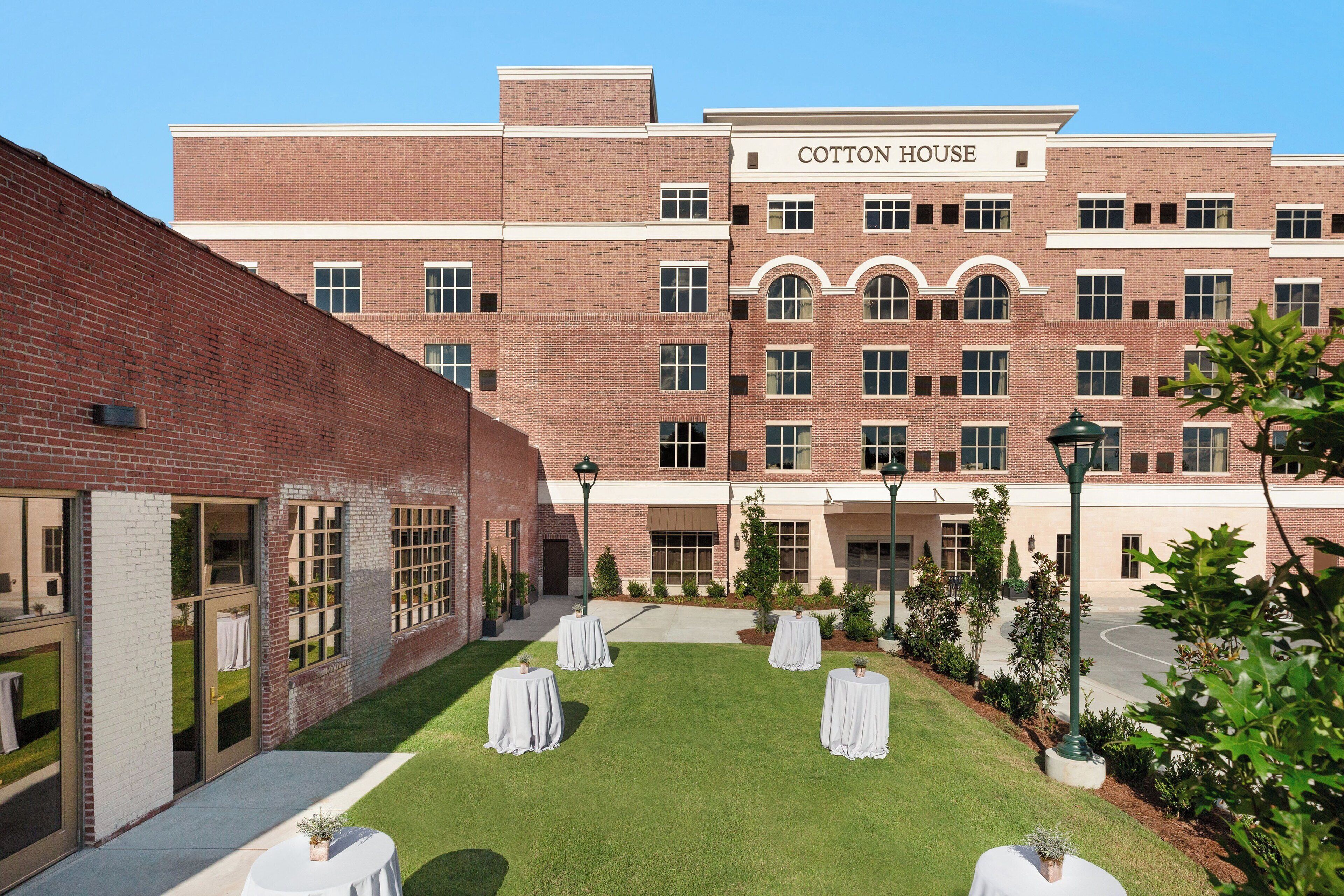 A brick building named Cotton House with a courtyard featuring four round tables with white tablecloths and potted plants.