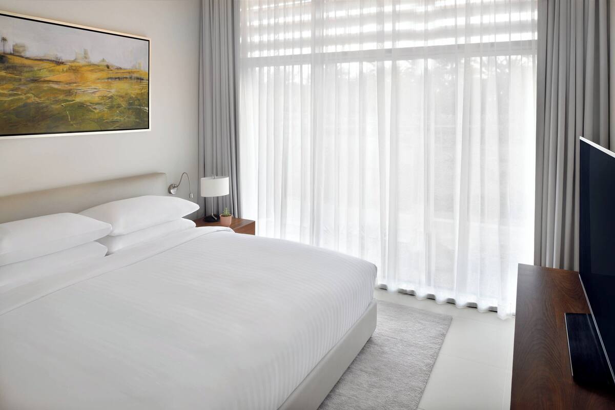 Ein Hotelzimmer mit einem großen Bett, einem Flachbildfernseher, einem Nachttisch mit Lampe und einem Bild an der Wand.