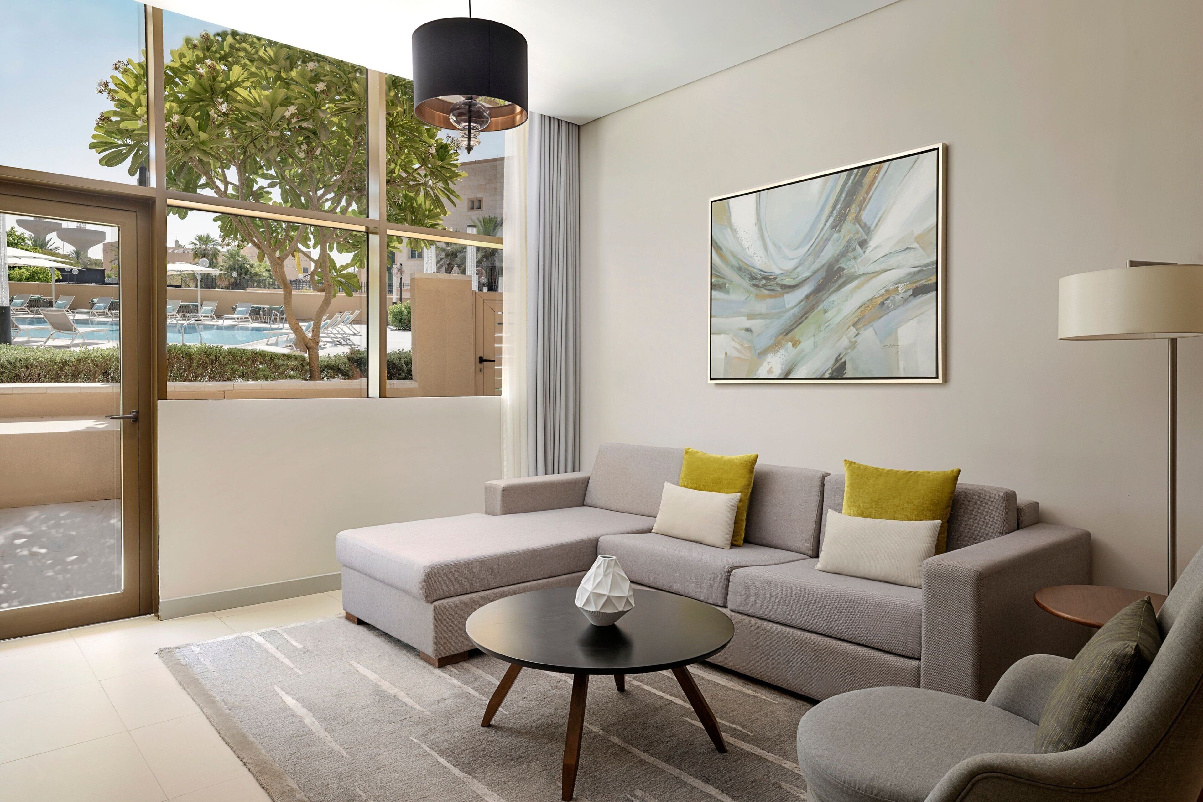 A modern living room with a grey sofa, a round coffee table, and a view of a pool through large windows.
