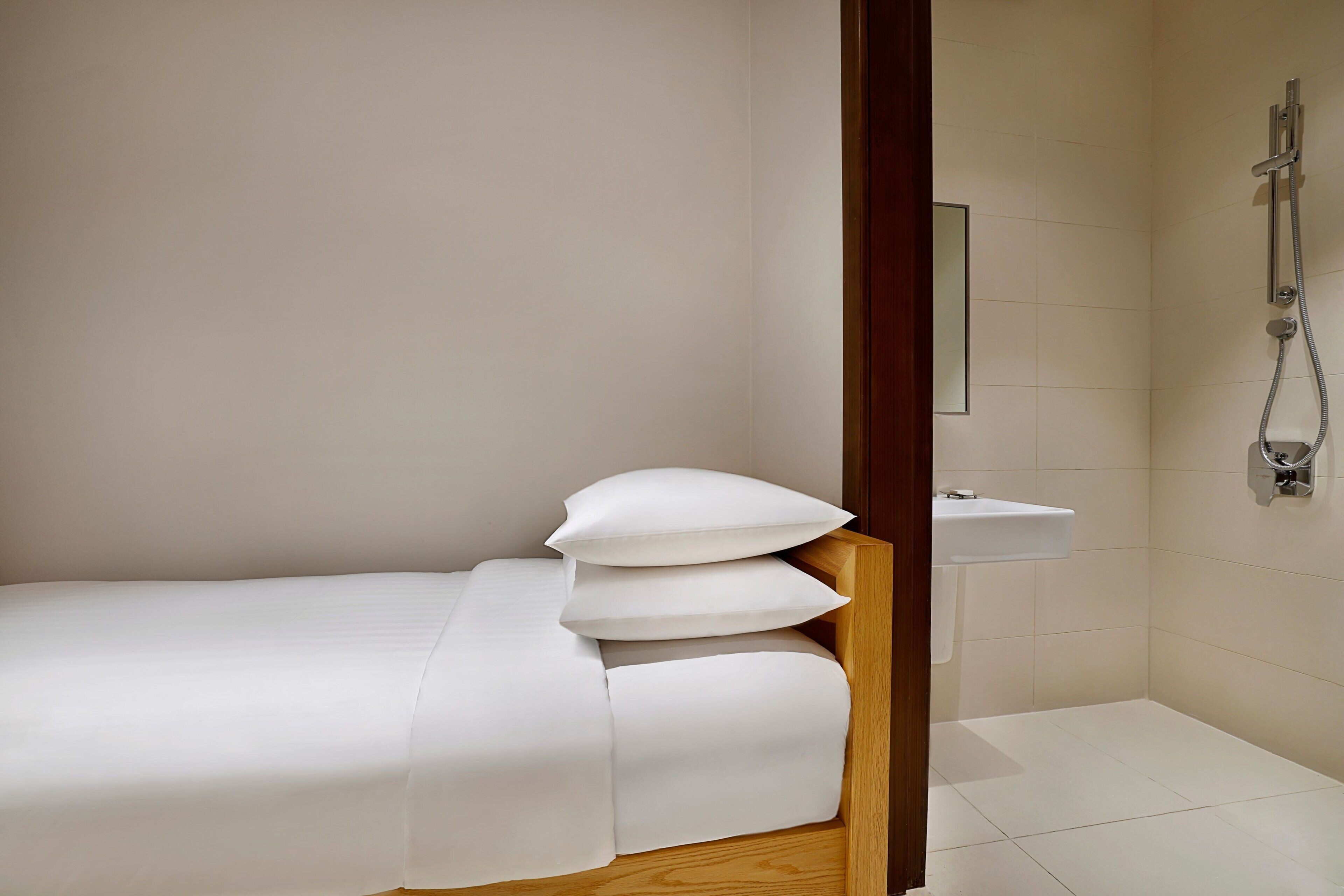 Un llit petit i ben fet amb roba de llit i almoades blanques, un capçal de fusta i un vàter blanc amb un raig de dutxa portàtil en una cantonada d'una habitació amb parets sense decoració i un paviment de rajola.