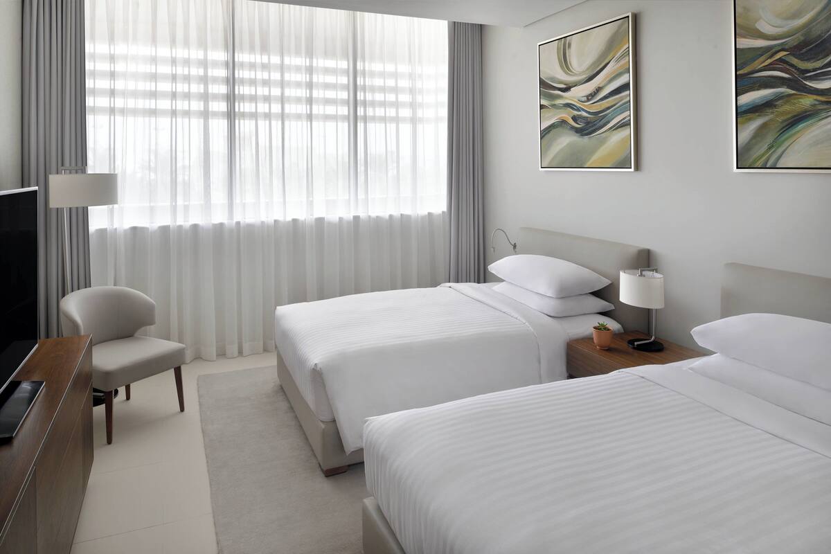 Ein Hotelzimmer mit zwei Betten, einem Schreibtisch, einem Sessel, einem Fernseher und Bildern an den Wänden.