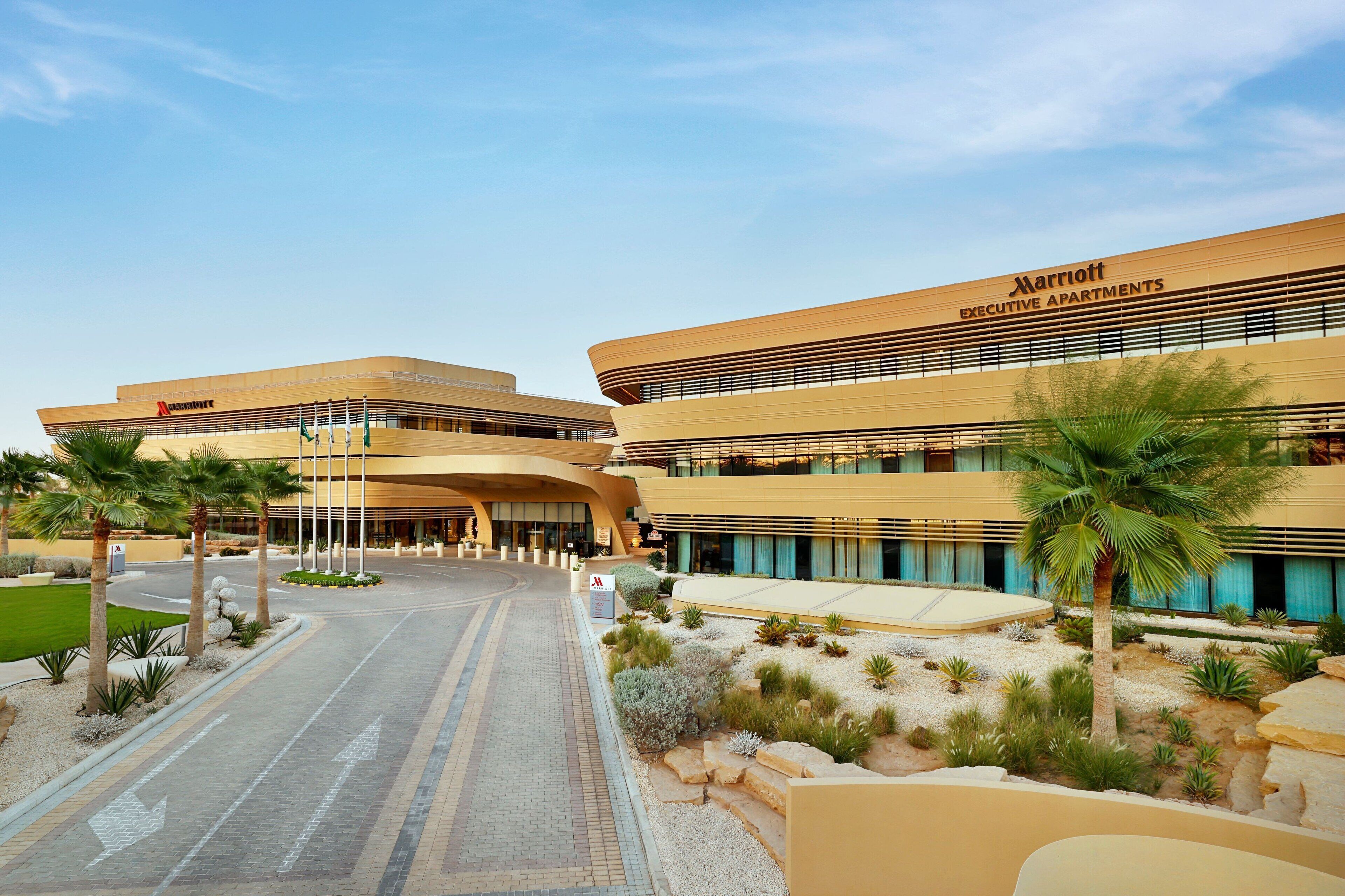 A modern hotel with a curved facade, multiple floors, and a landscaped entrance area with palm trees and shrubs.