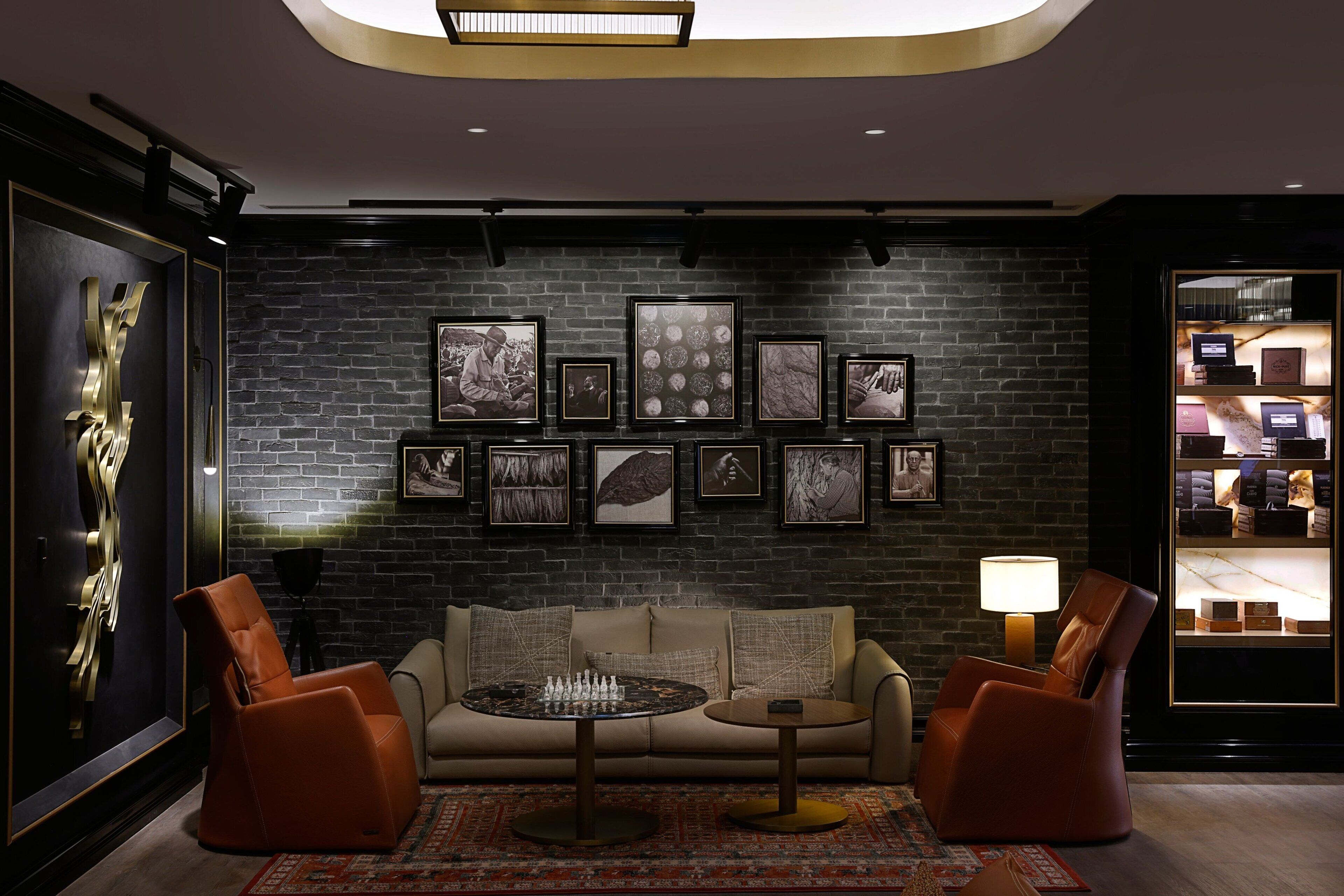 A modern lounge with a dark brick wall, a sofa, two armchairs, a round coffee table, and a wall-mounted display of framed pictures.