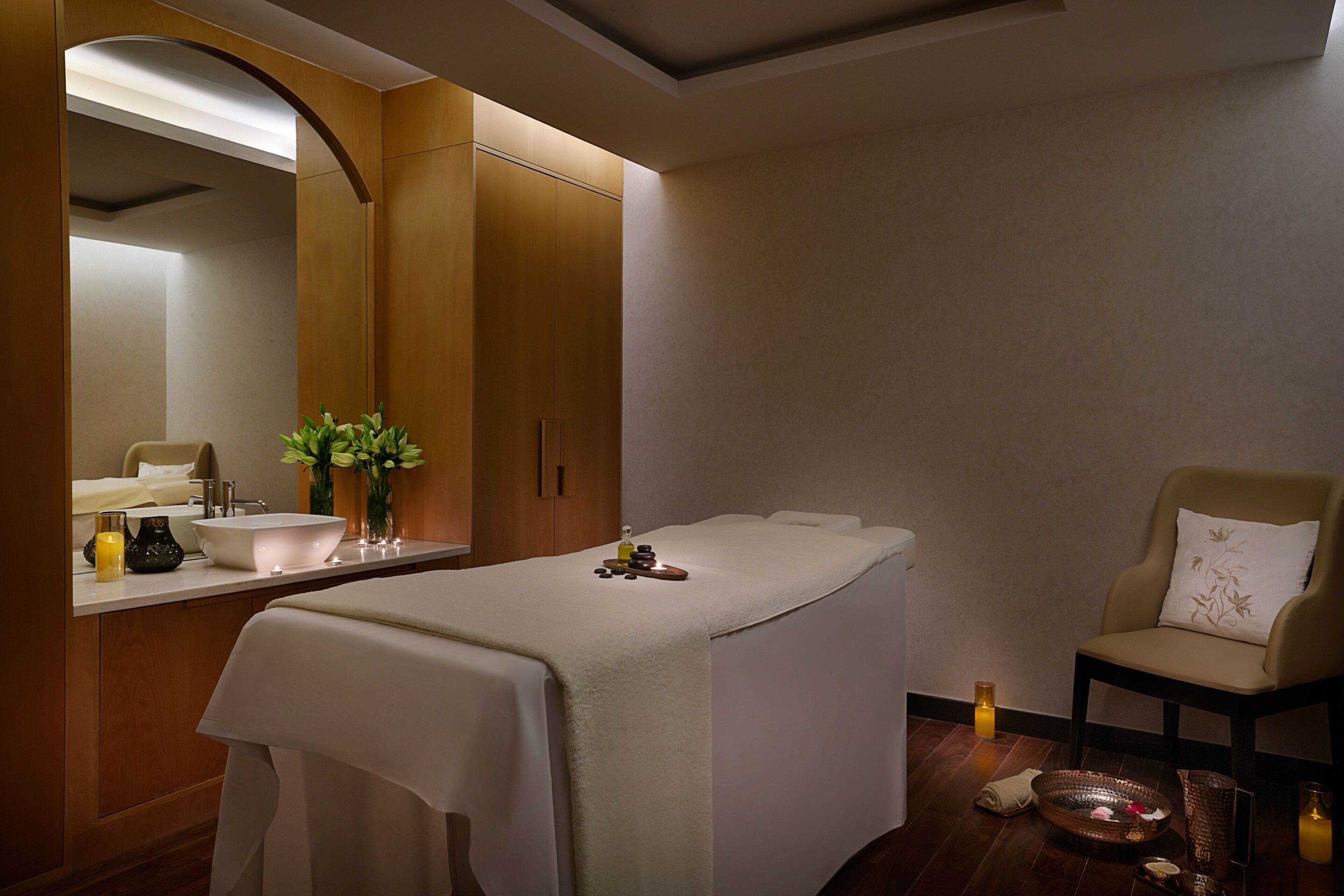 A spa room with a massage table, a chair, a sink, and a mirror.