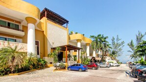 Un hotel con fachada amarilla, columnas blancas y un área de entrada cubierta. Se ven palmeras y un cielo azul despejado.