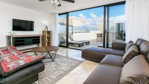 A modern living room with a flat-screen TV, a ceiling fan, a sofa, a coffee table, and a balcony with outdoor seating. - Penthouse A-Nah by Stella Rentals (Playa del Carmen)