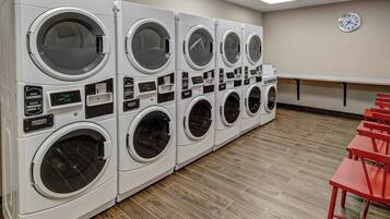 A row of white front-loading washers with control panels, a clock on the wall, and a red bench in a laundry room.
