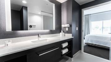 Un baño moderno con un espejo grande, lavamanos blanco y muebles oscuros. Hay un perchero y una ventana con vistas a la ciudad.