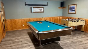 Une table de billard, avec une table de baby-foot à côté, des lambris en bois sur les murs et deux tableaux encadrés accrochés au mur.