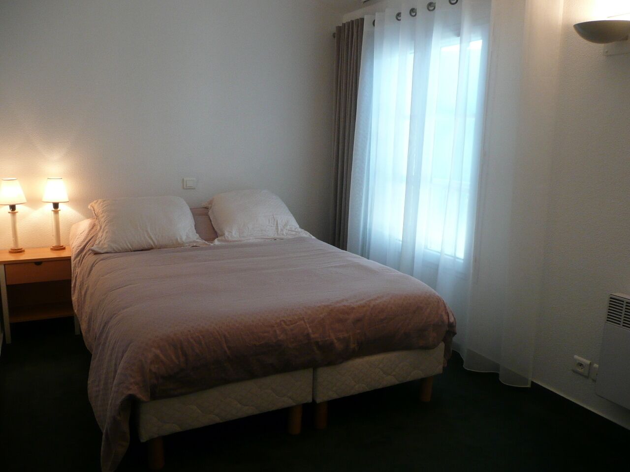 Een eenpersoonsbed met een beige sprei en kussens, een nachtkastje met twee lampen en een raam met dunne gordijnen.