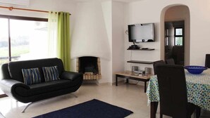 A living room with a black sofa, a television, a fireplace, a dining table with chairs, and a large window with curtains. - 2 Bedroom Apartment with Swimming Pool at São Rafael Beach - Albufeira (Albufeira)