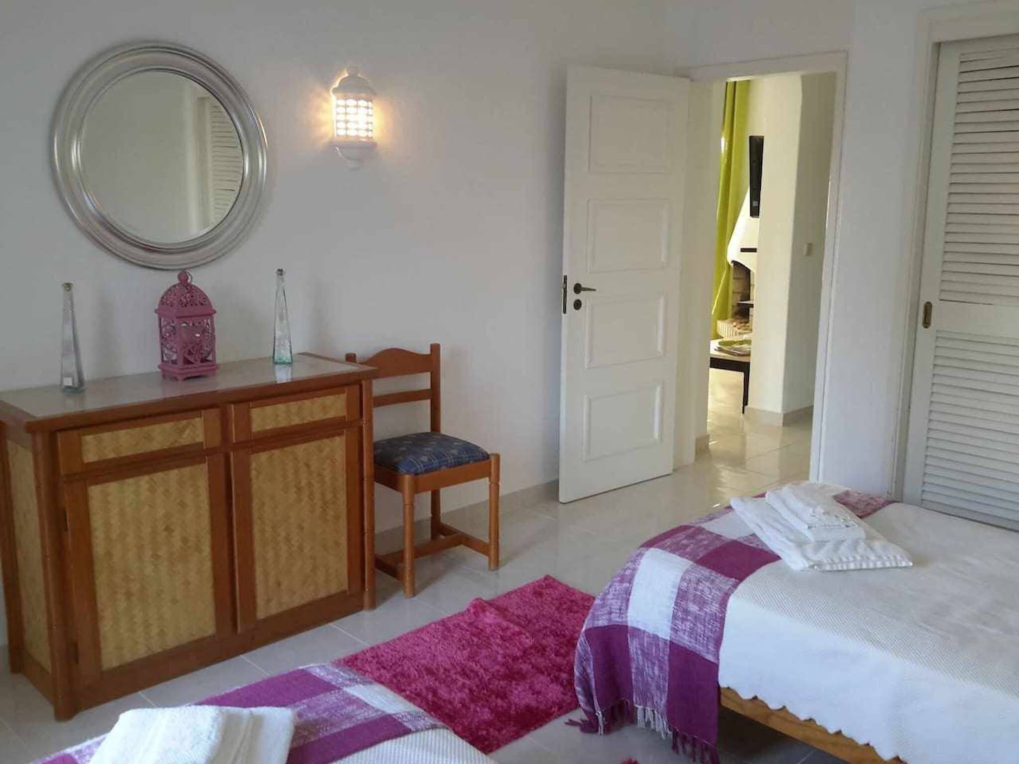 A bedroom with a bed, a desk with a chair, a mirror, and a door leading to another room.