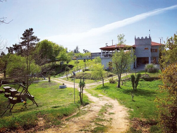 A rustic building with a red-tiled roof, surrounded by a grassy area with a dirt path leading to a picnic table and swing set.