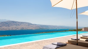 An infinity pool with a view of the sea and mountains, lounge chairs with towels, and a pergola with a large umbrella.