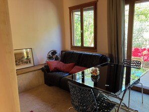 Un espace de vie comprenant un canapé en cuir noir, une table en verre, un ventilateur et une vue sur la verdure environnante.