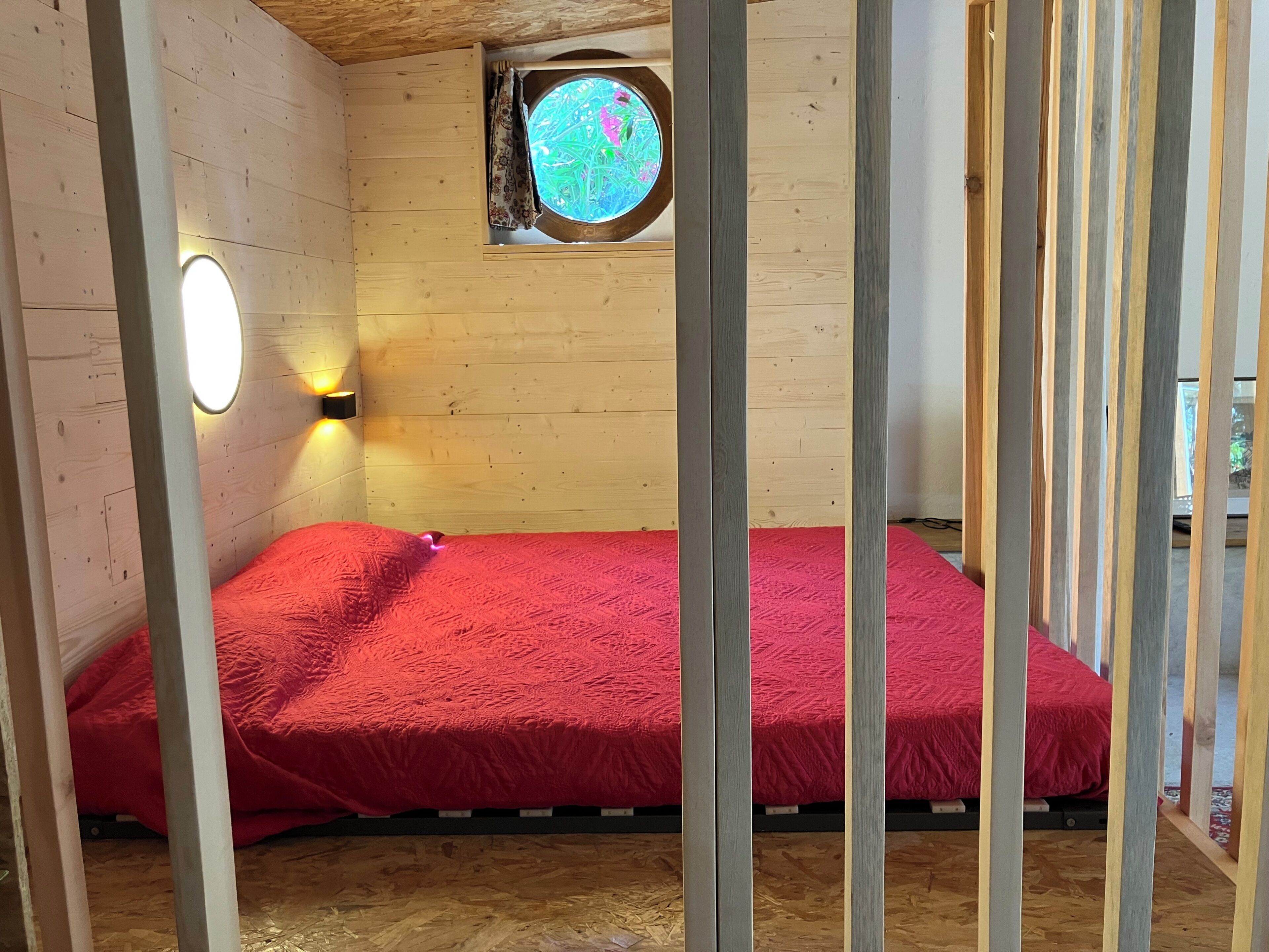 A room with a wooden floor, a bed with a red blanket, a round window with a view, and a wall-mounted light.