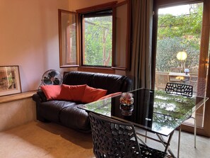 Un espace de vie comprenant une table en verre, un canapé en cuir avec des coussins rouges, un ventilateur et une vue sur la verdure environnante.